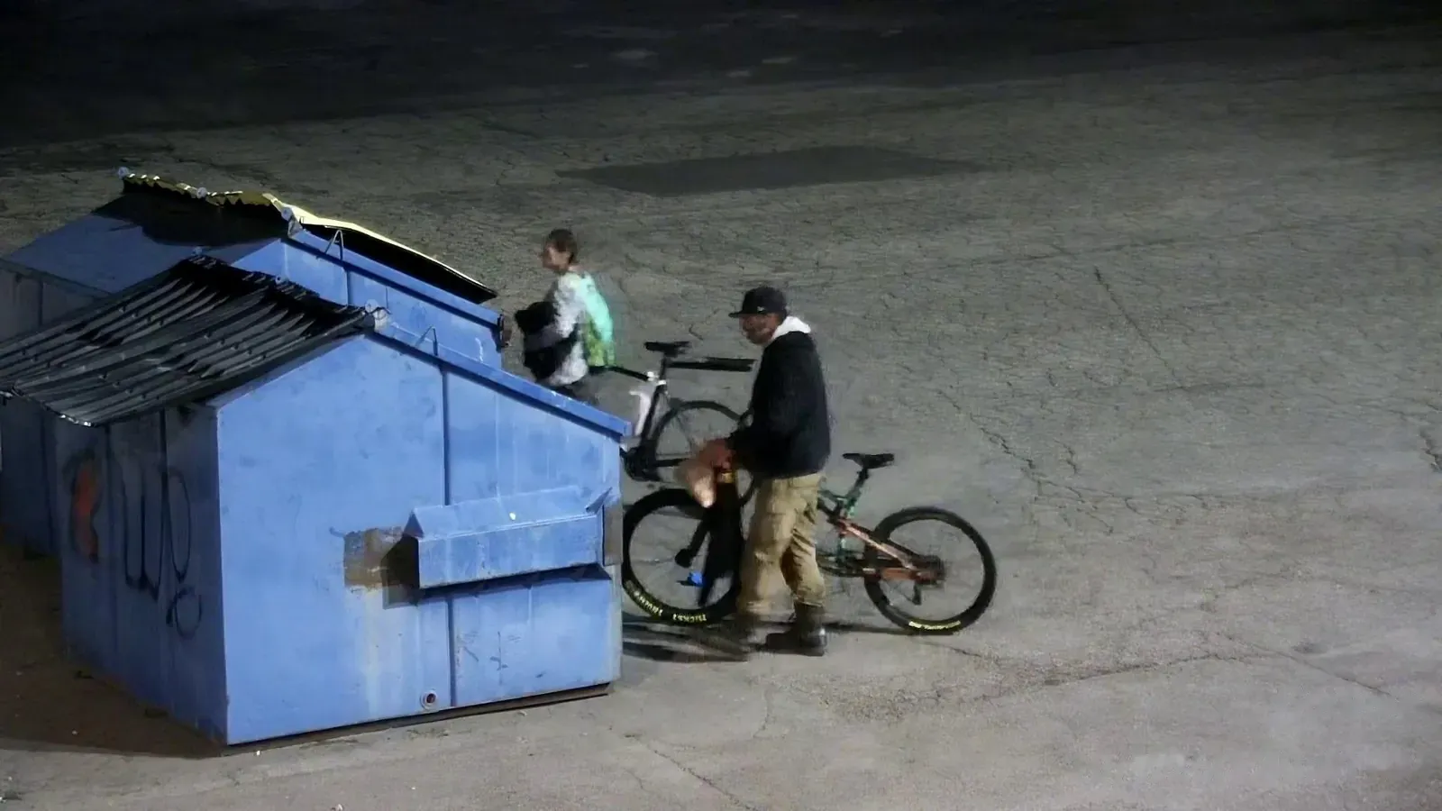 Two people and a bicycle by a dumpster at night